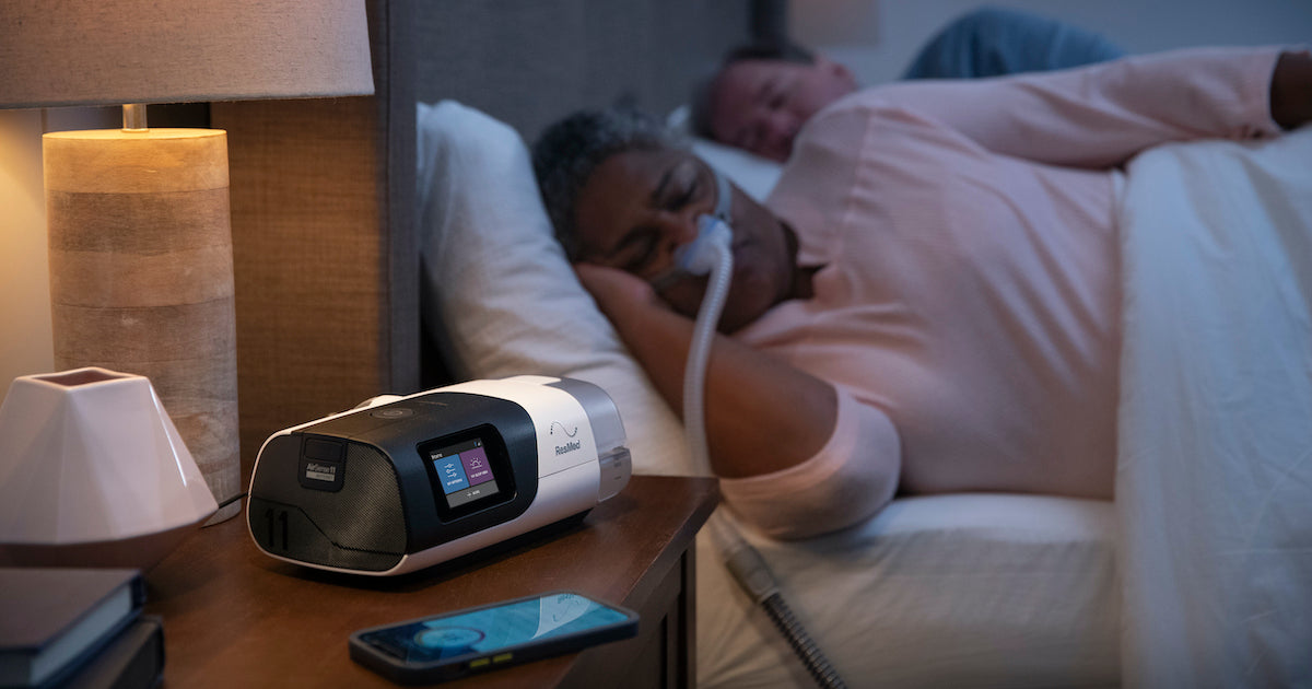 AirSense 11 AutoSet CPAP machine placed on a nightstand beside a woman sleeping in bed with a nasal CPAP mask. The CPAP machine is sleek and modern, with a digital display showing therapy settings. The device is connected to the water chamber and hose.