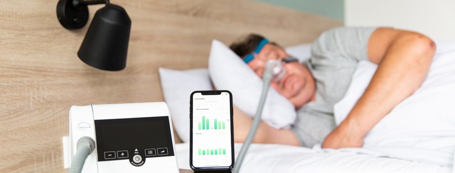 Löwenstein CPAP/APAP device on a bedside table next to a cellphone displaying the integrated app, with the hose connected to a mask worn by a man sleeping in bed.