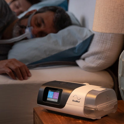 AirSense 11 AutoSet CPAP machine is placed on a nightstand beside a person sleeping in bed with a nasal CPAP mask. The CPAP machine is sleek and modern, with a digital display showing therapy settings. The device is connected to the water chamber and hose.