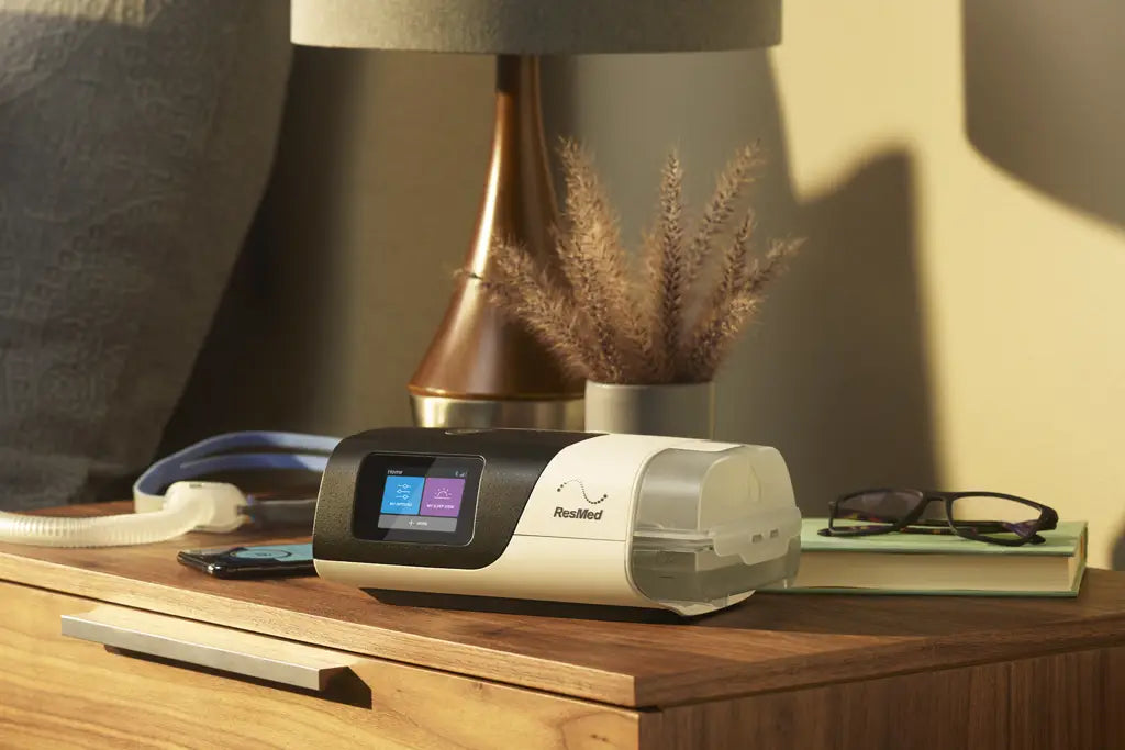 A sleek and modern AirSense 11 AutoSet CPAP machine sits on a nightstand, next to a pair of glasses resting on a book, with a nasal pillows mask on the opposite side. The machine features a digital display showing therapy settings and is connected to a water chamber.